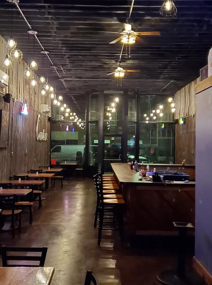 An interior view of a restaurant with wooden tables and chairs, decorative lighting, and a bar area.