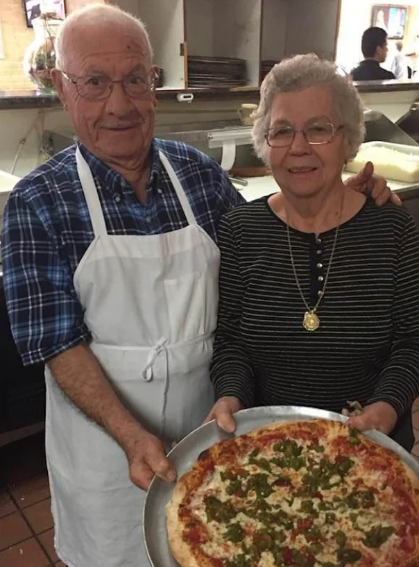 Couple proudly holding a fresh Margherita pizza in a cozy pizzeria
