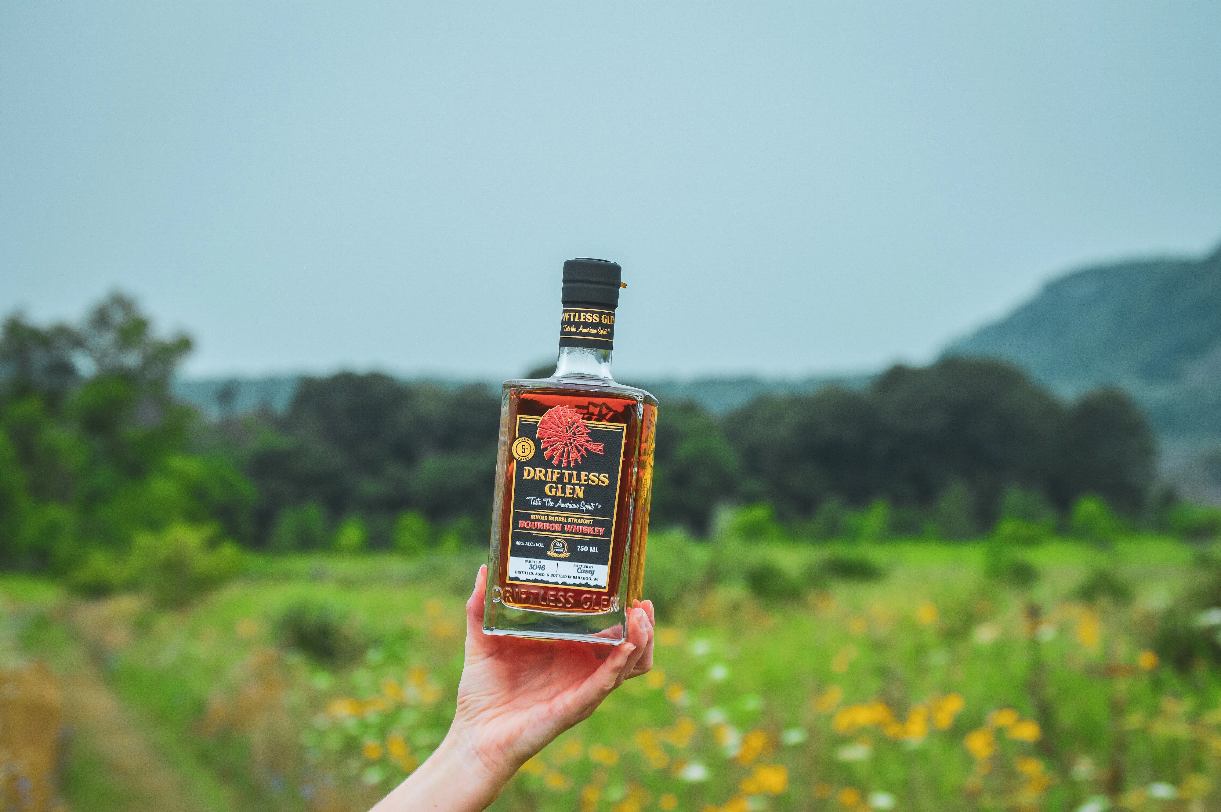 A hand holds up a full bottle of Single Barrel Bourbon outside in a field with flowers in the background