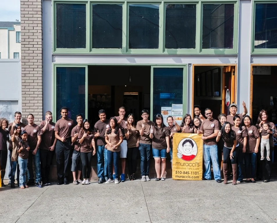 Muracci's staff gathered in front of the restaurant, posing together in matching branded T-shirts while holding a Muracci's sign.