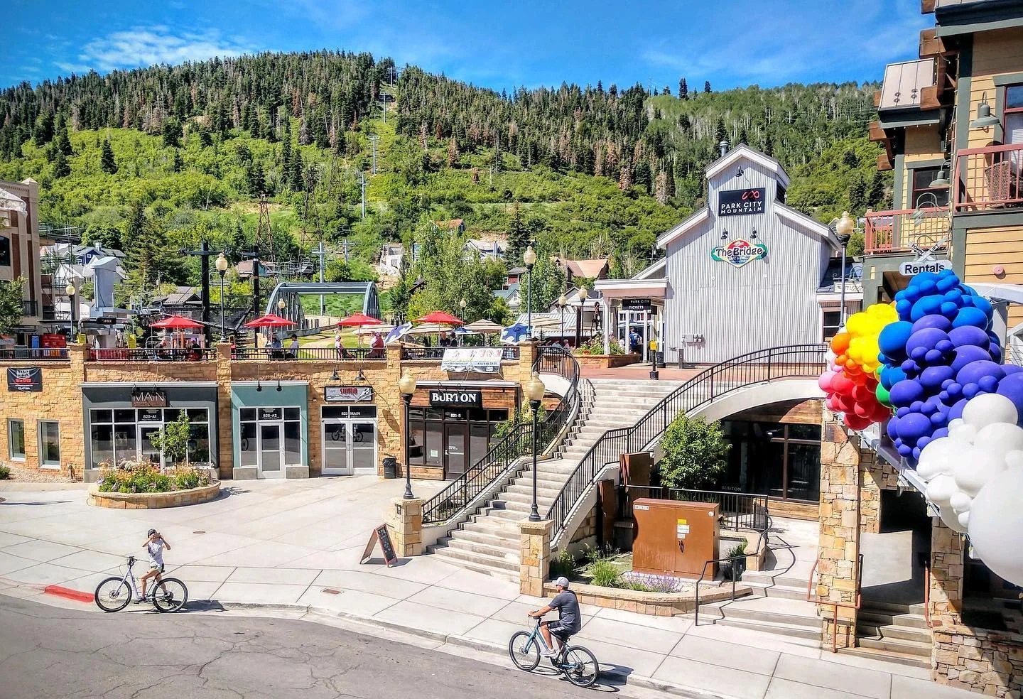 A lively outdoor shopping area featuring colorful umbrellas, bike paths, and a backdrop of green hills.