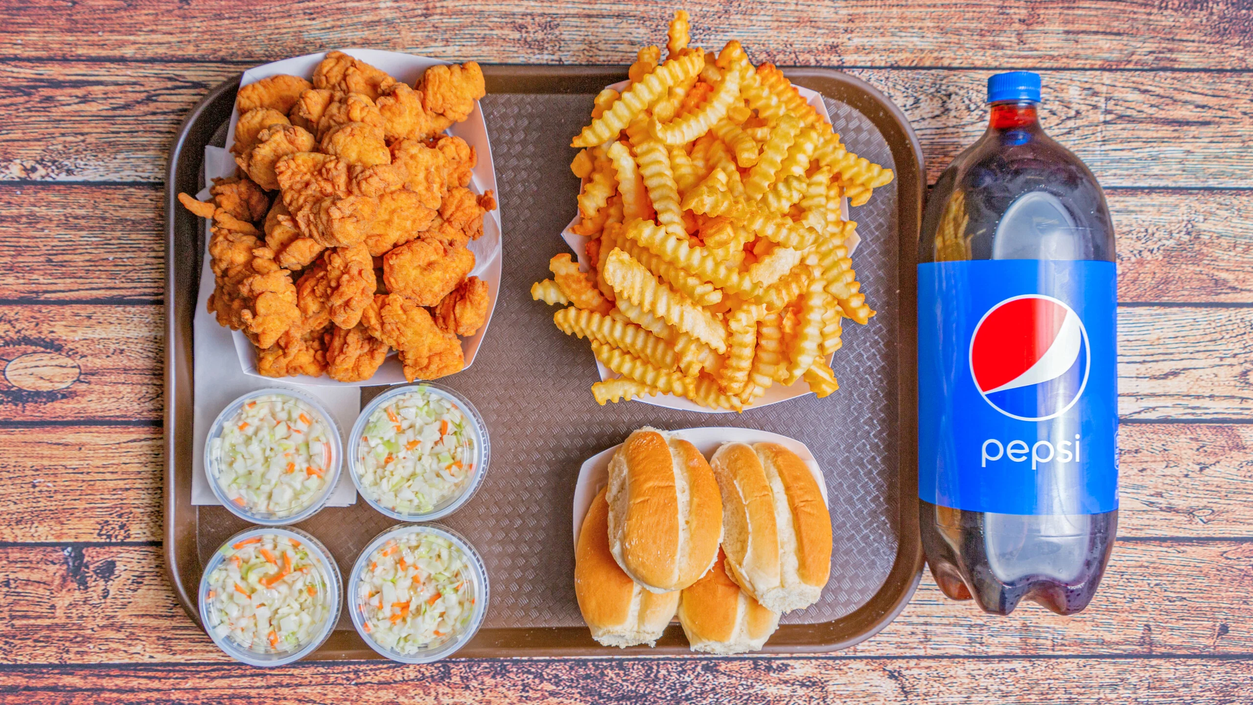 A serving tray filled with fried chicken pieces, crinkle-cut fries, coleslaw in cups, and a bottle of Pepsi.