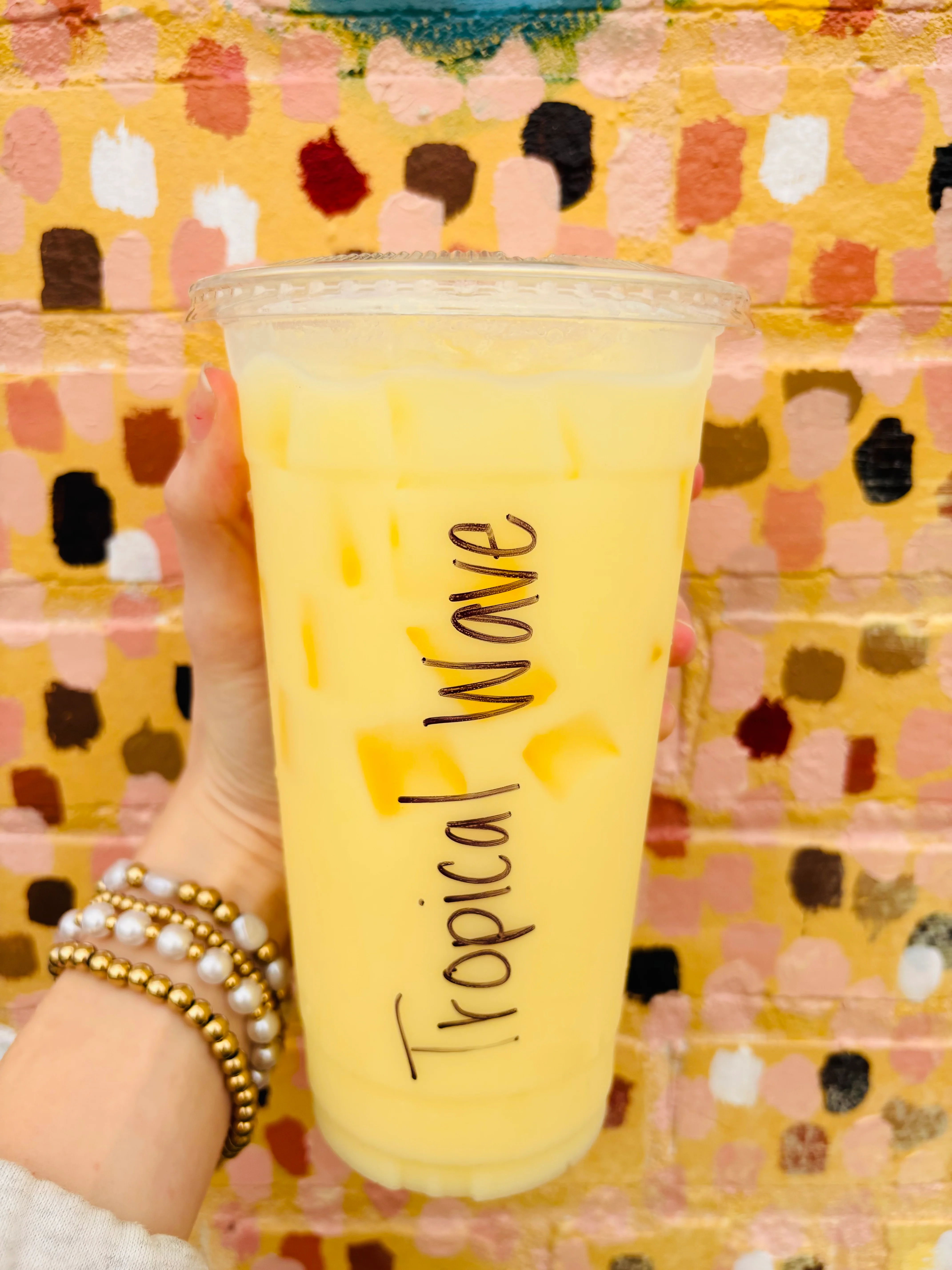 A large cup of yellow drink with ice, labeled as 'Tropical Wave', held against a colorful, patterned background.