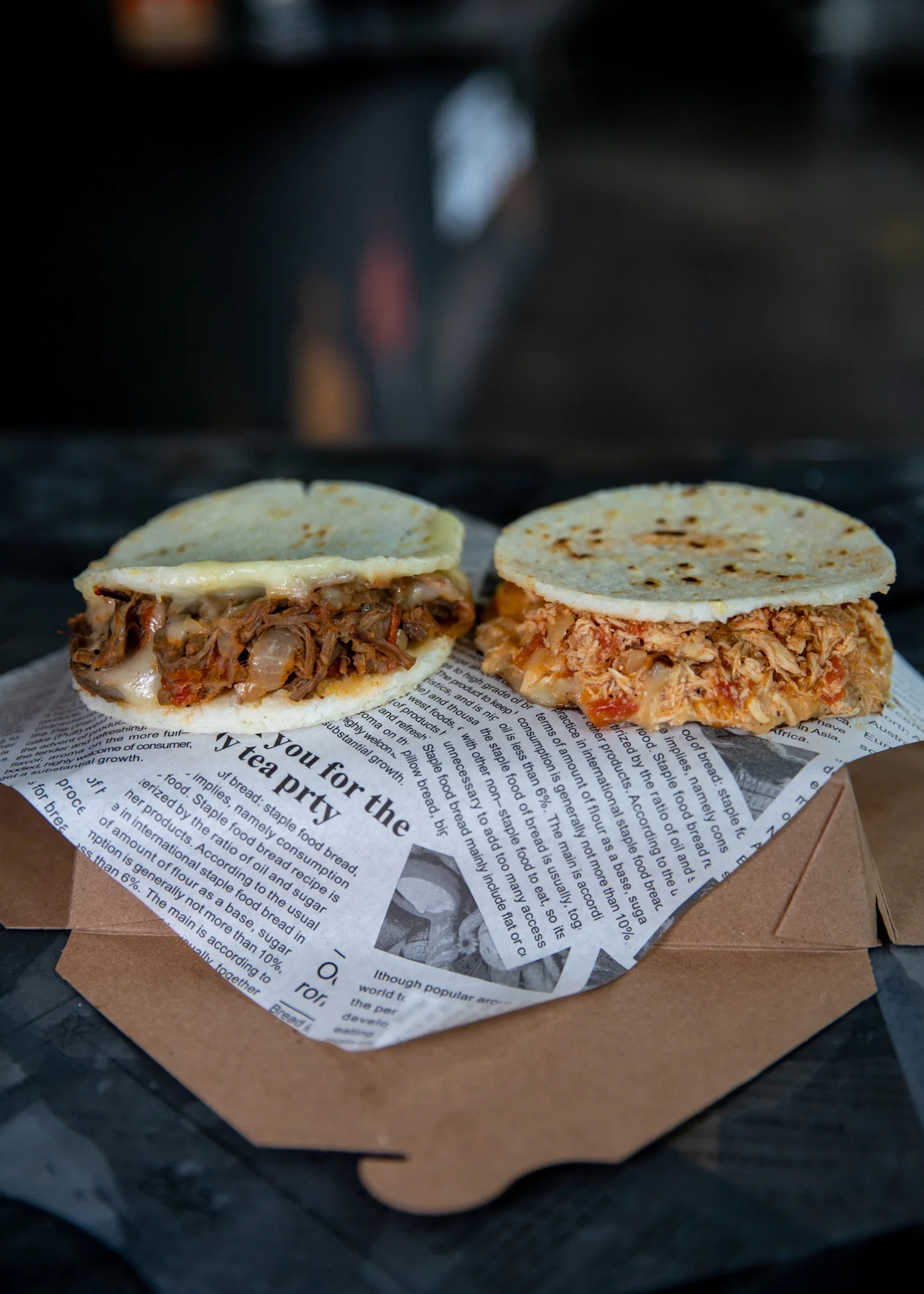 Two arepas filled with various meats, cheese, and sauces, served on a bed of newspaper.
