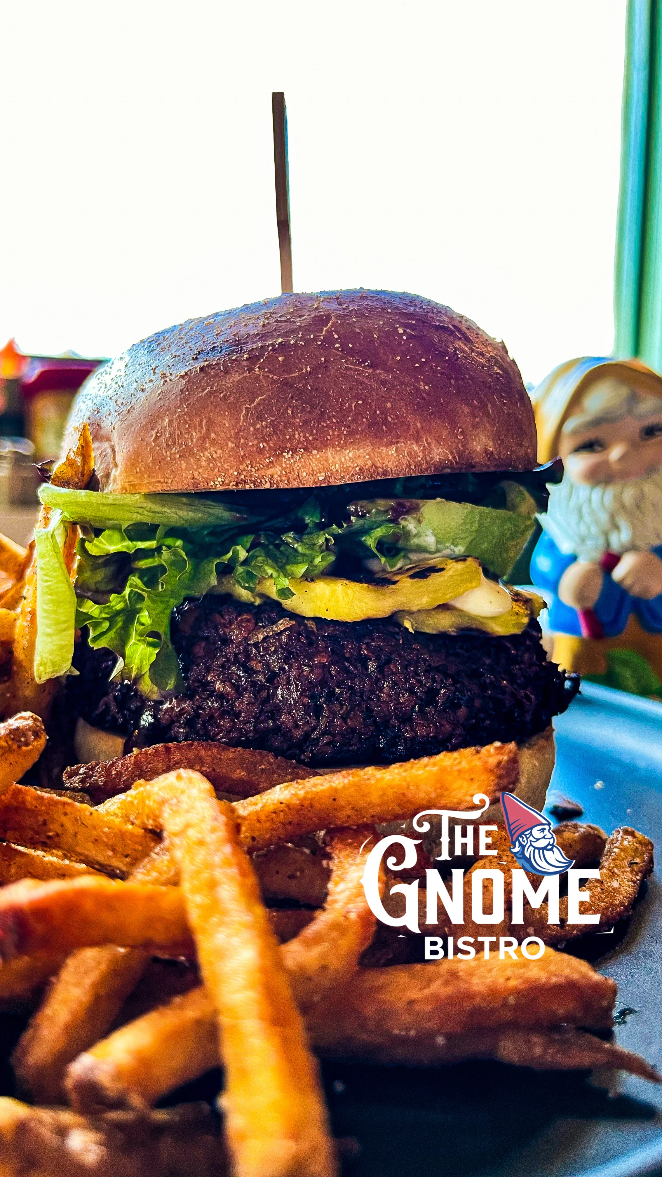 A gourmet burger topped with lettuce, pickles, and a slice of cheese, served with a side of crispy fries. The meal is presented on a plate with a colorful background.