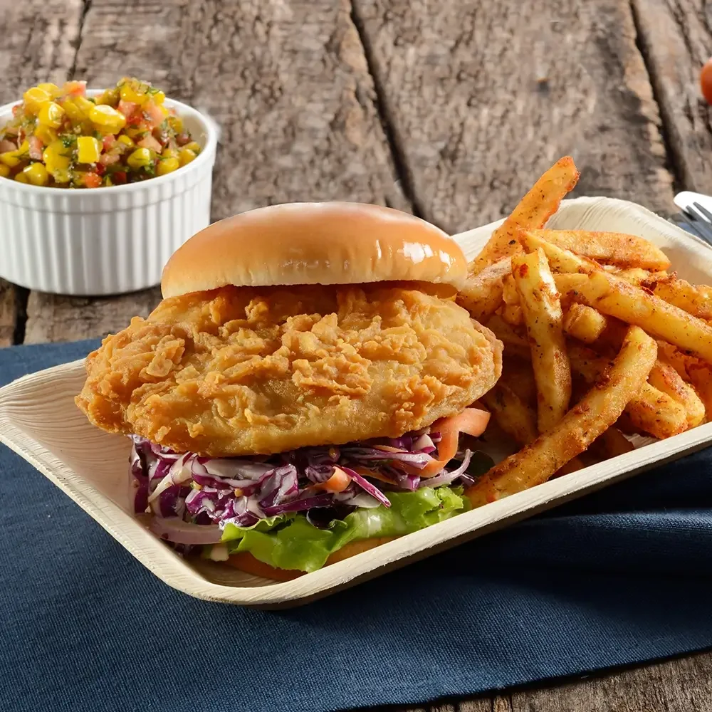 A crispy fried chicken sandwich with coleslaw and tomato, accompanied by seasoned fries and a side of salsa.