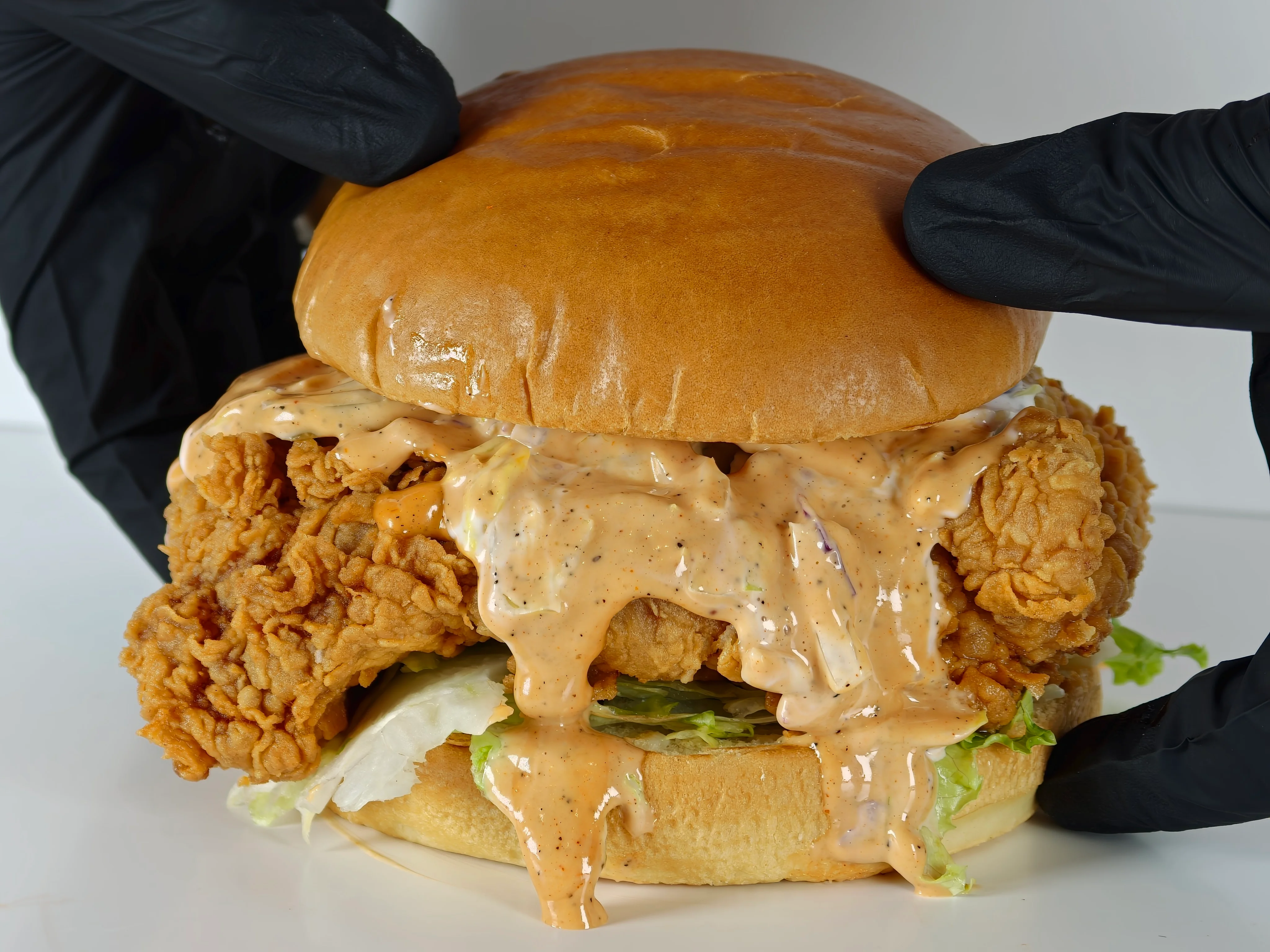 A close-up of a chicken sandwich loaded with crispy fried chicken, lettuce, and a creamy sauce, held by a hand in a black glove.