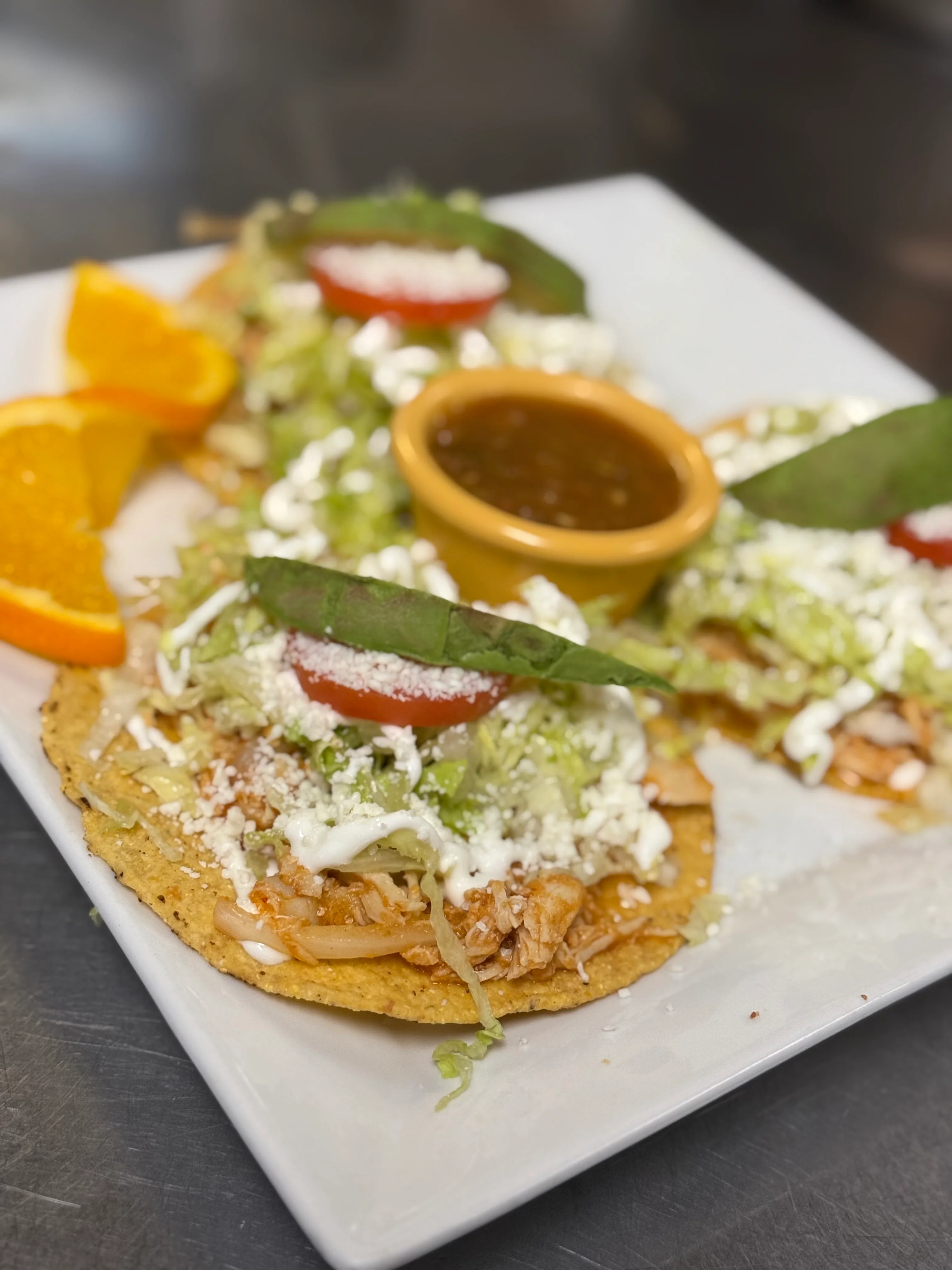 A plate featuring three tacos topped with shredded meat, lettuce, tomato, cheese, and avocado leaves, accompanied by a small bowl of salsa and orange wedges.