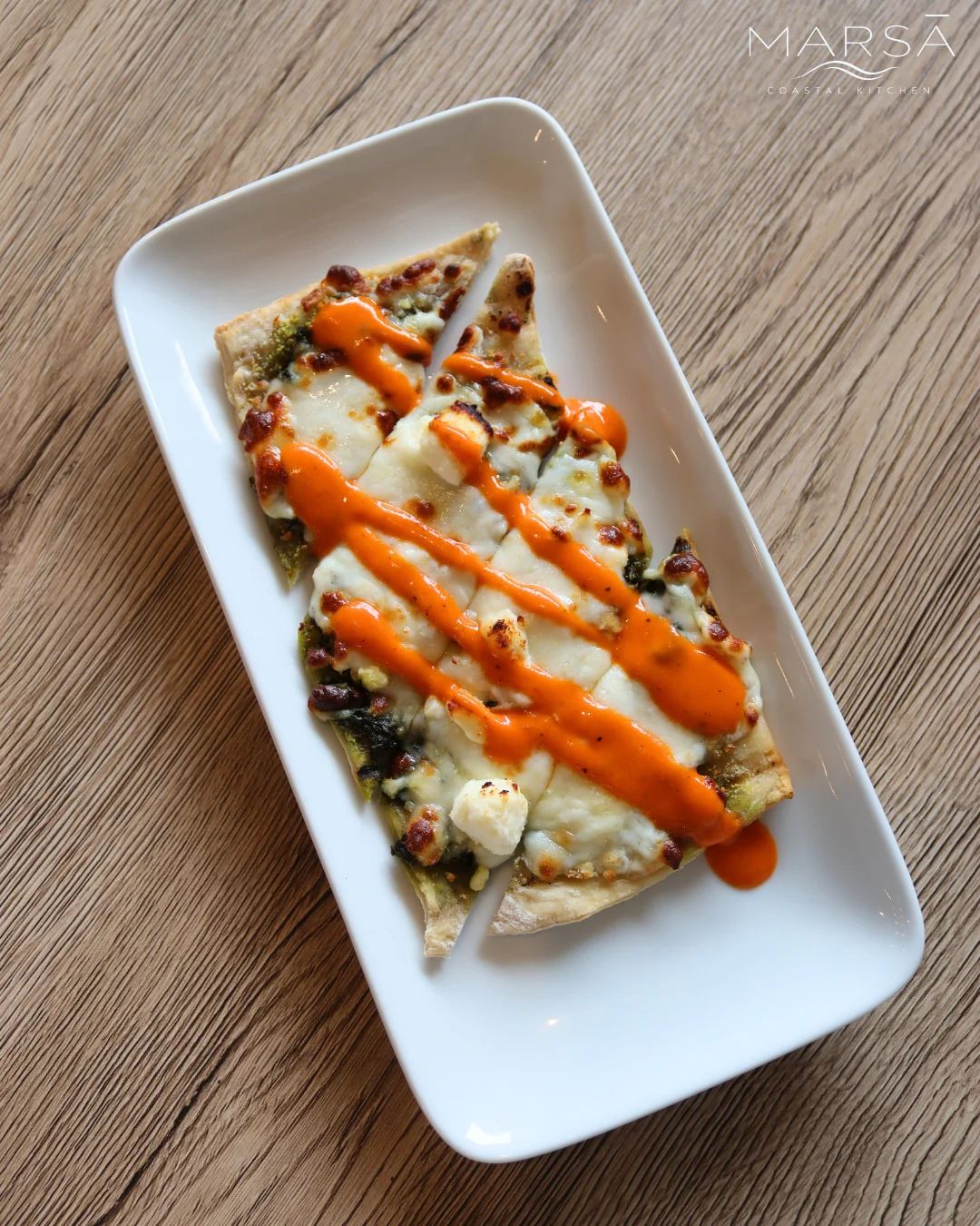 A slice of flatbread topped with cheese and drizzled with a spicy sauce, presented on a rectangular plate.