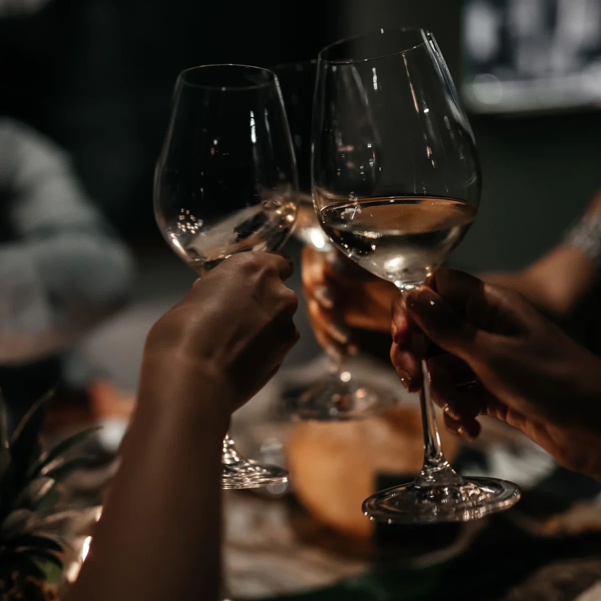 Three people clink glasses of white wine in a celebratory toast at a dimly lit table.