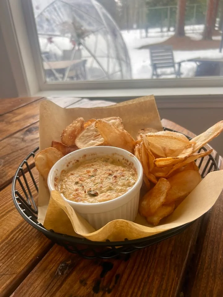 A basket containing a creamy dip and a side of crisp potato chips, placed on a wooden table with a snowy outdoor view in the background.