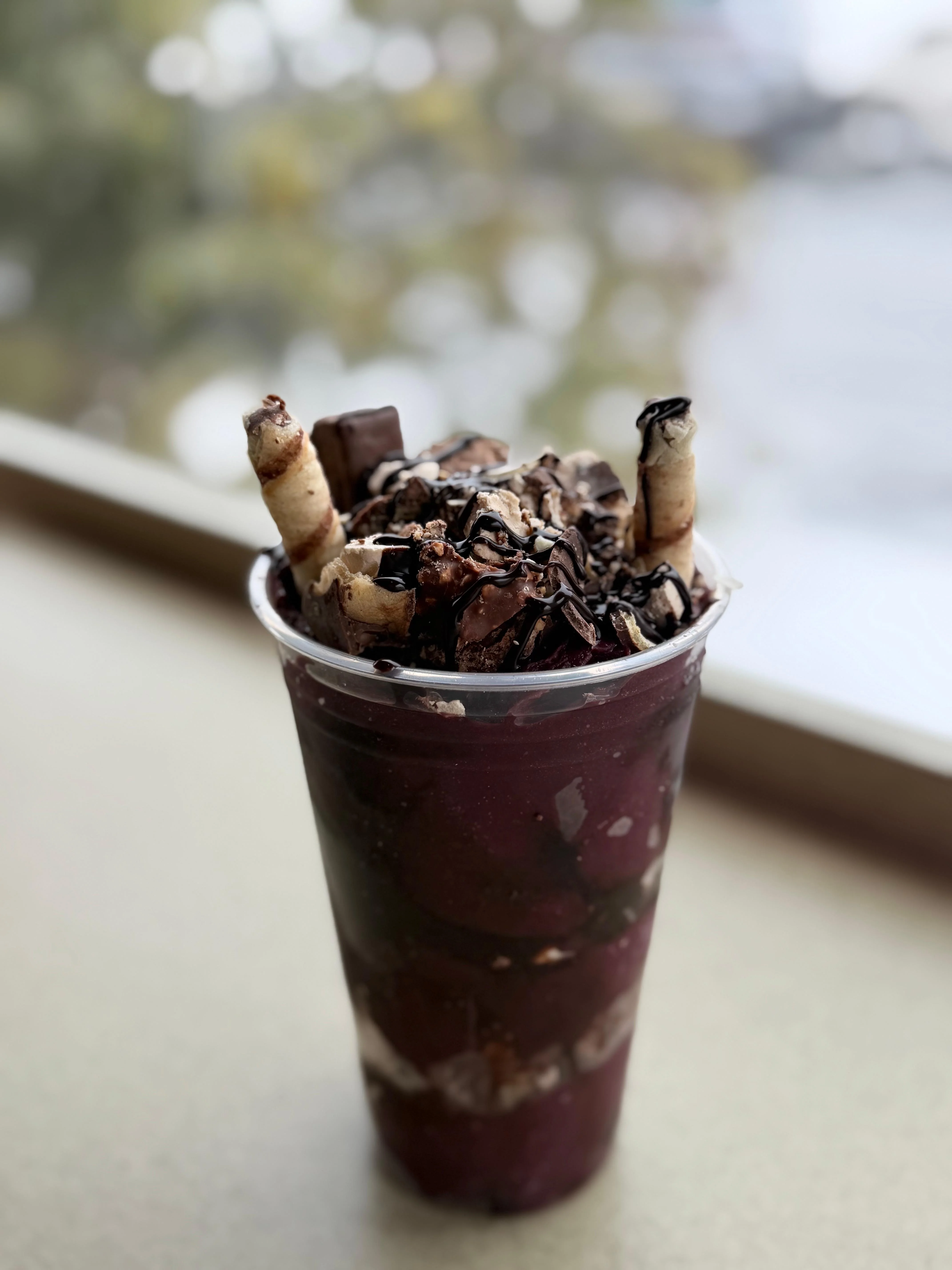 A layered dessert in a clear cup featuring dark purple açaí, topped with chocolate shavings and rolled wafer sticks.
