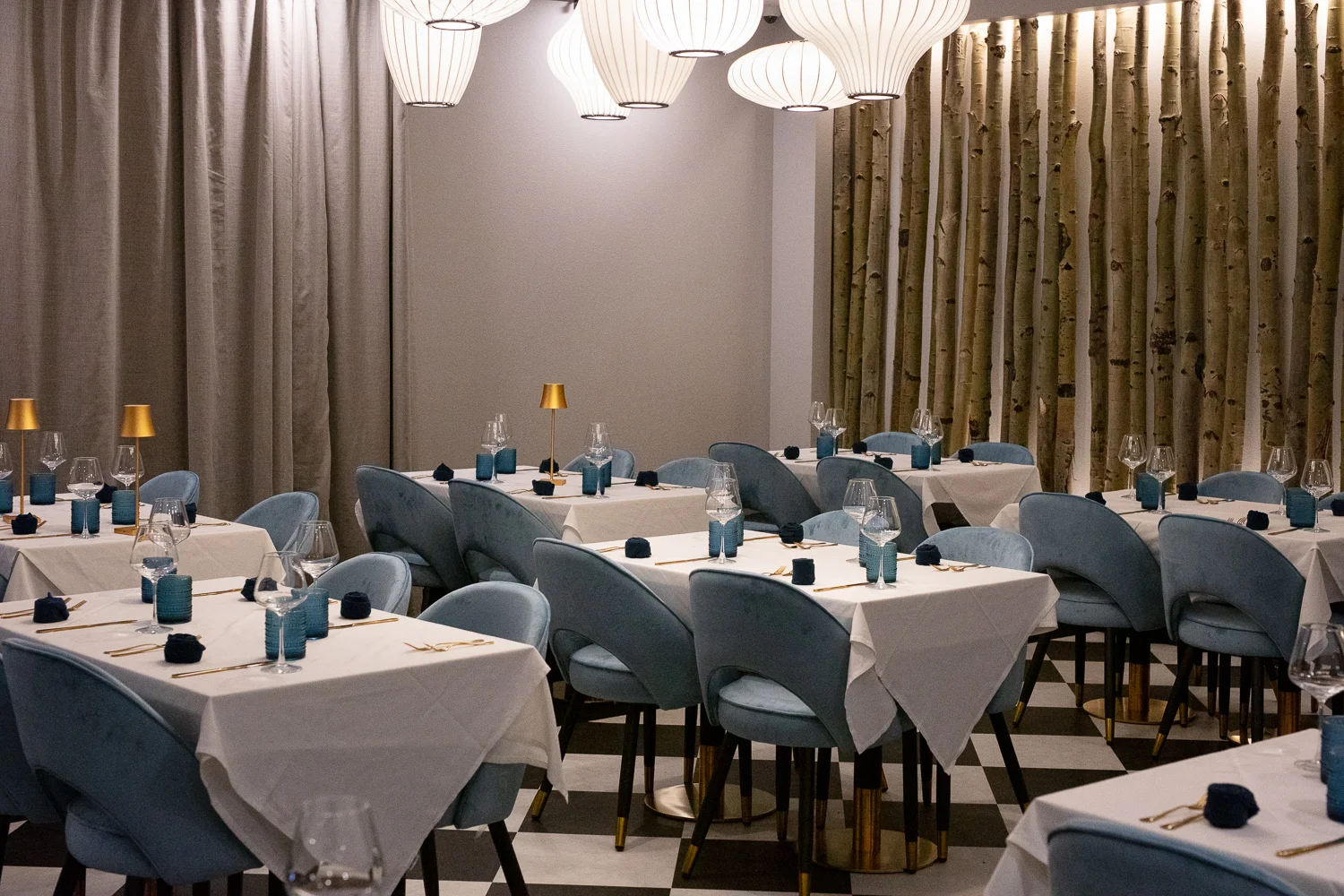 Elegant restaurant with soft blue velvet chairs, white tablecloths, and gold accents. Sculptural pendant lights hang above, with birch trunks and gray curtains adding a natural, modern touch.