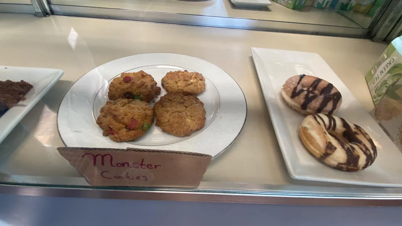 A display of baked goods including two monster cookies and two donuts. The cookies are chunky with visible ingredients and colorful bits, while the donuts have a decorative icing and chocolate drizzle.