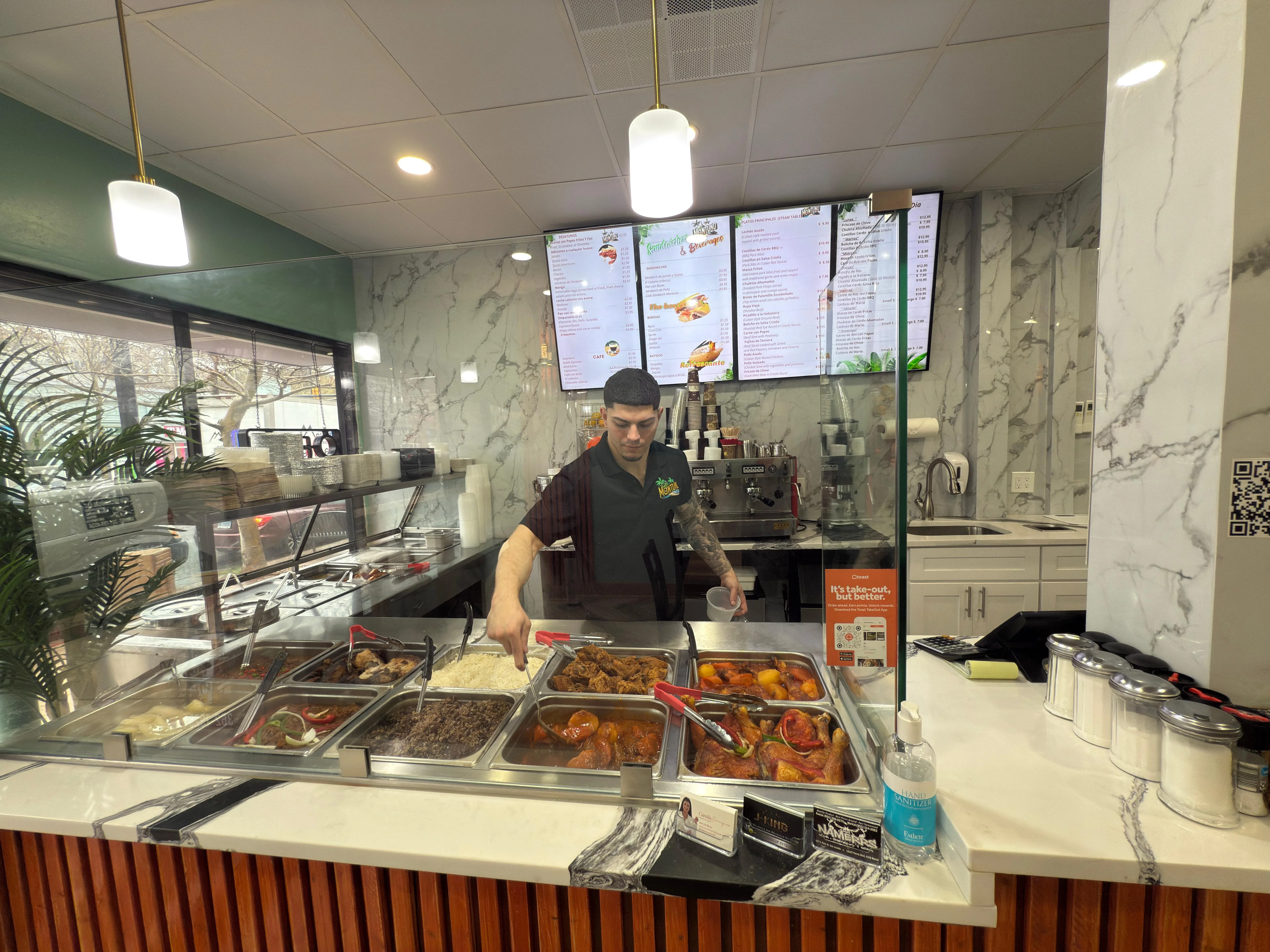 Food service counter in a restaurant with trays of rice, beans, and cooked dishes. Above are digital menu screens displaying items; the interior has modern marble accents.