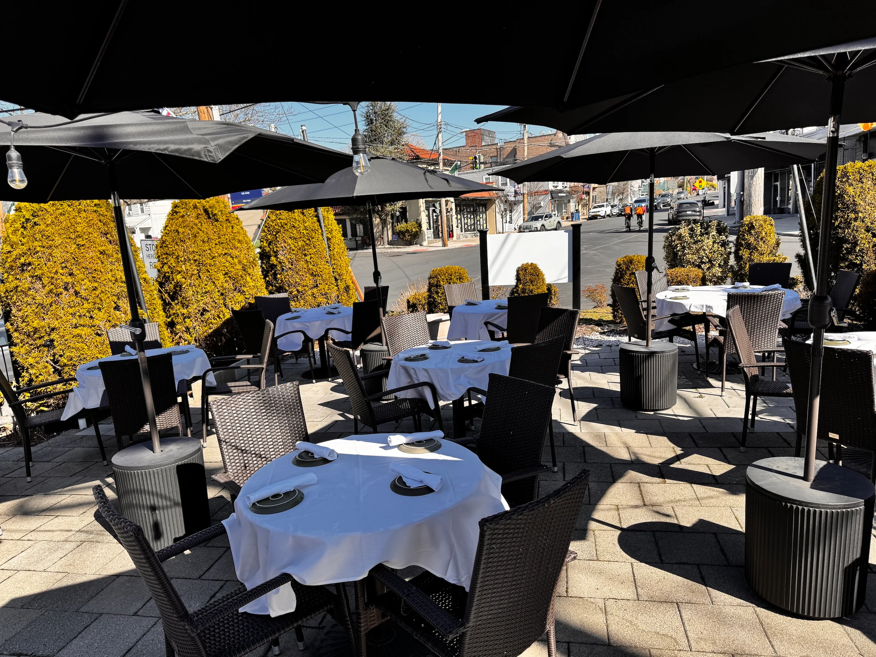 Outdoor restaurant patio with round tables and black woven chairs, covered by large umbrellas. Sunny day with a relaxed, inviting atmosphere.