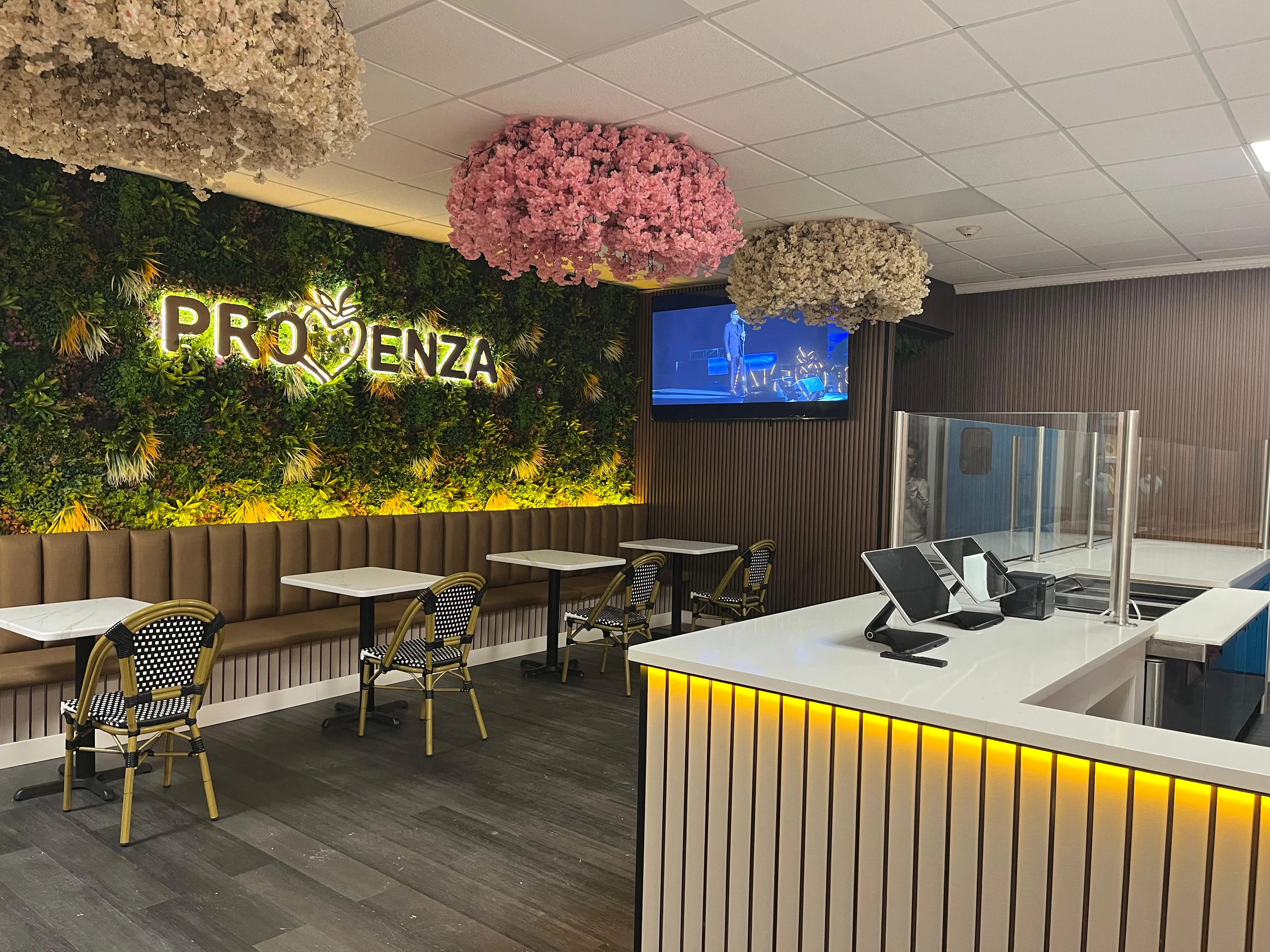 A modern cafe interior featuring a lush green wall, decorative flowers above, and a sleek counter with digital ordering screens.
