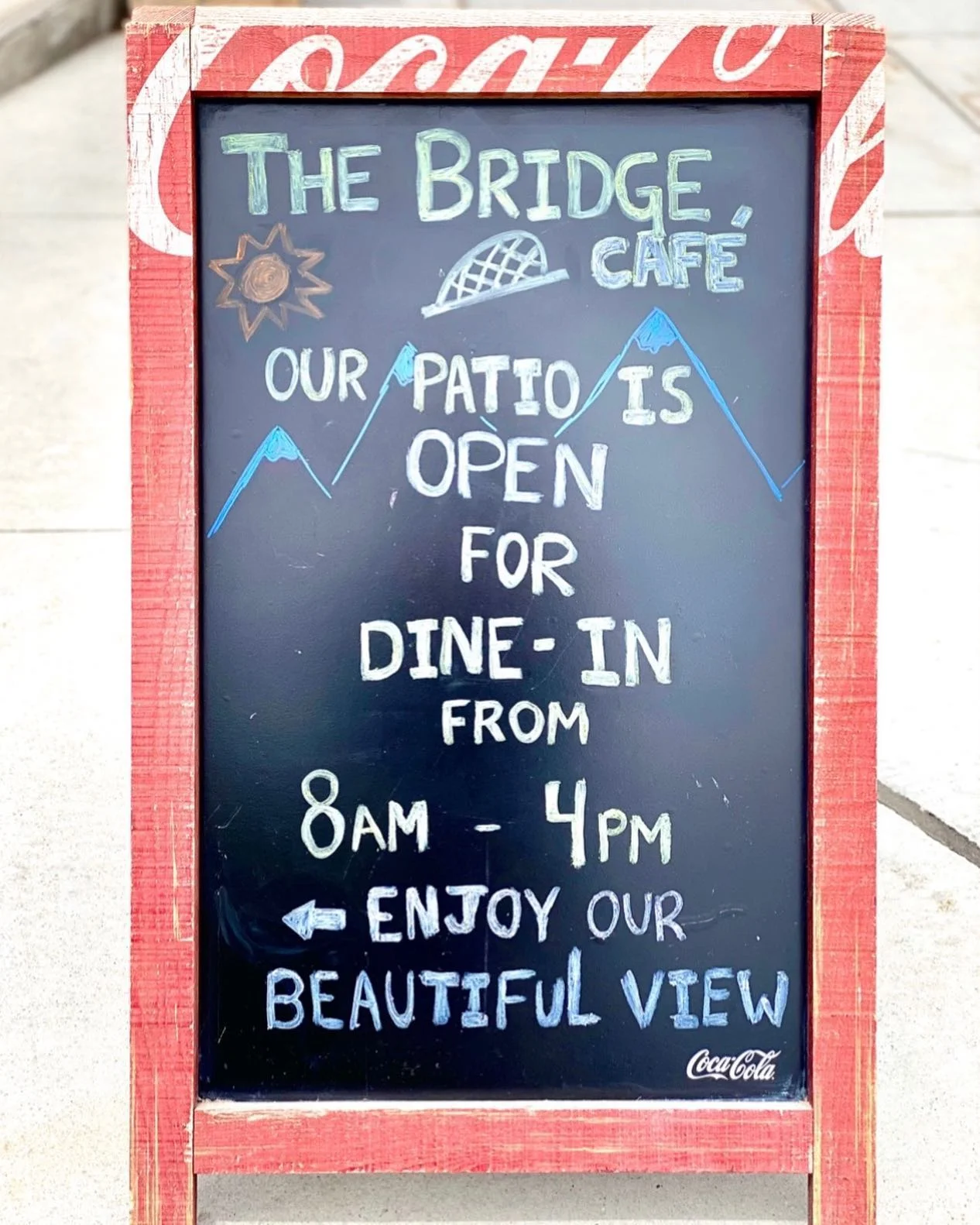 A chalkboard sign announcing the opening hours of a café patio, inviting customers to enjoy a beautiful view while dining.