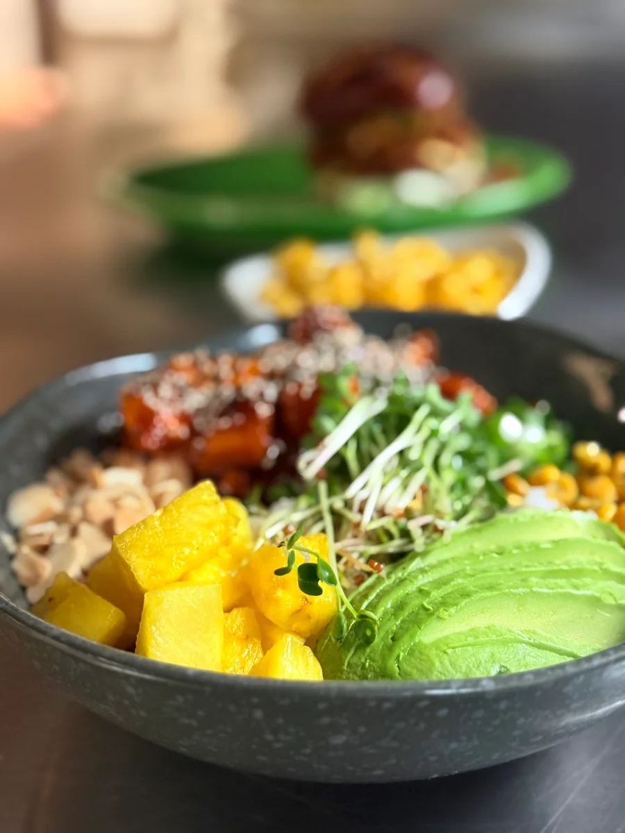 A colorful Buddha bowl featuring rice topped with sauced sweet potatoes, sesame seeds, greens, corn, avocado slices, pineapple chunks, and sliced almonds.