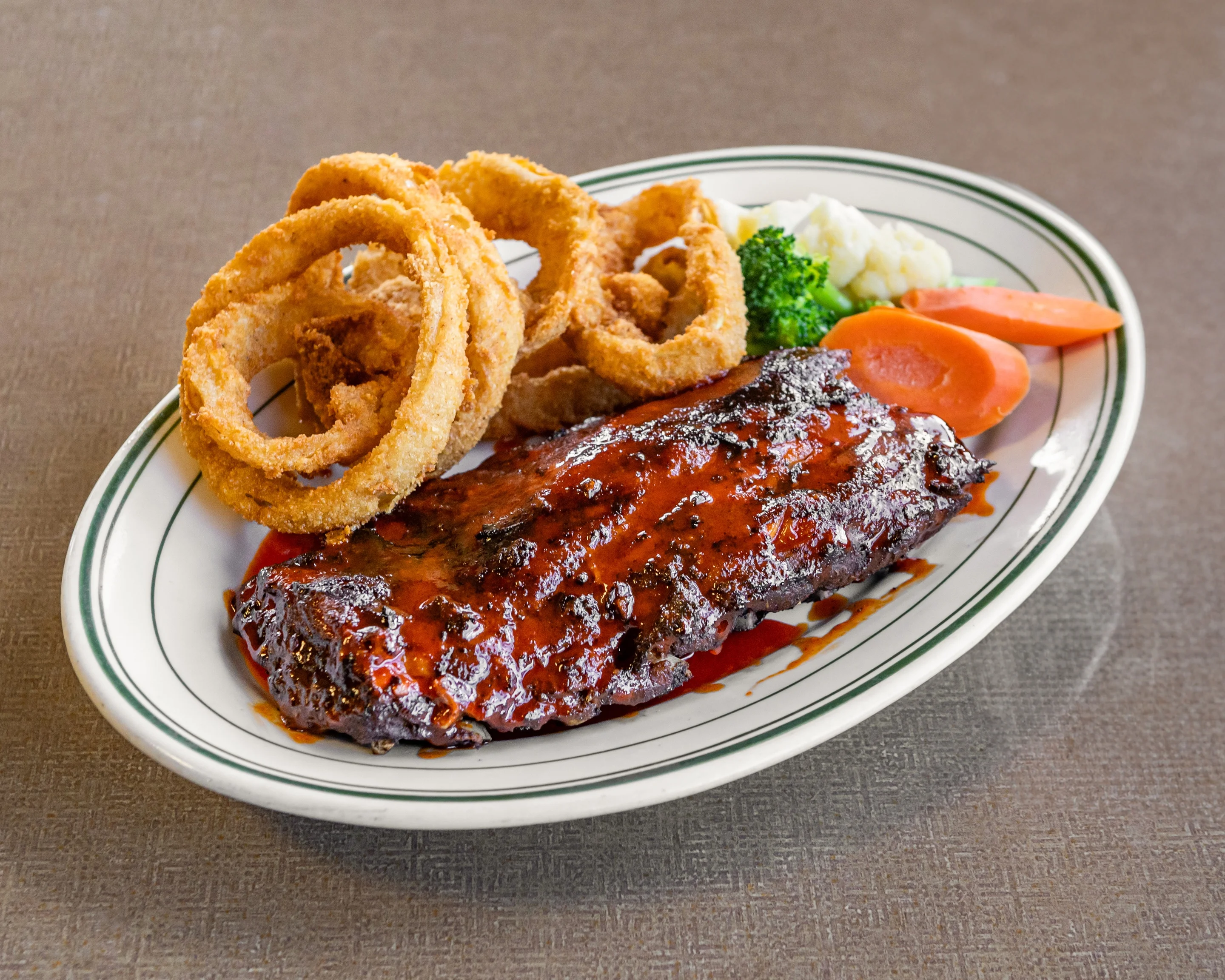 A barbecue rib meal featuring a glazed rib, onion rings, and steamed vegetables, served on an oval plate.