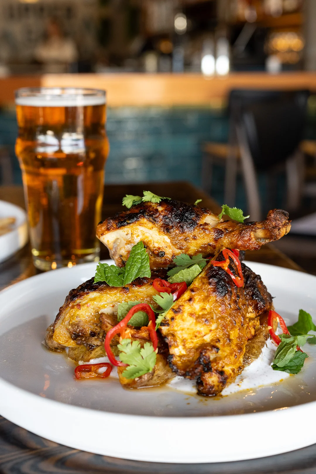 A plate of grilled chicken garnished with herbs and sliced red chili, accompanied by a glass of beer in the background.