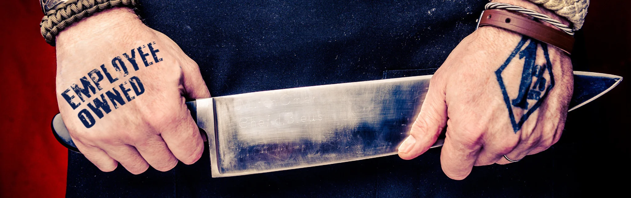 Chef holding a knife with 'Employee Owned' tattoo on hand