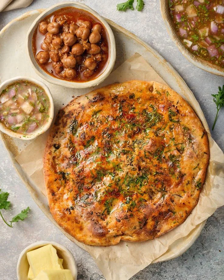 A platter featuring a crispy flatbread topped with herbs, accompanied by bowls of chickpeas and a tangy sauce. The dishes are arranged on a light-colored background.