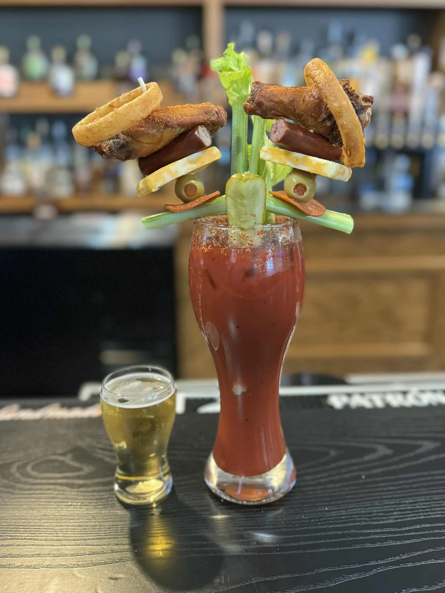 A Bloody Mary cocktail topped with celery, bacon, ham cubes, cheese, a boiled egg, and vegetables, with colorful slot machines in the background.