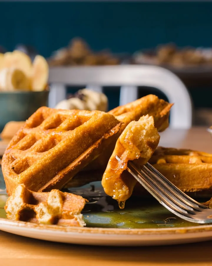 A plate of waffles drizzled with syrup, with one piece partially cut, revealing a fluffy interior. The scene is set on a wooden table.