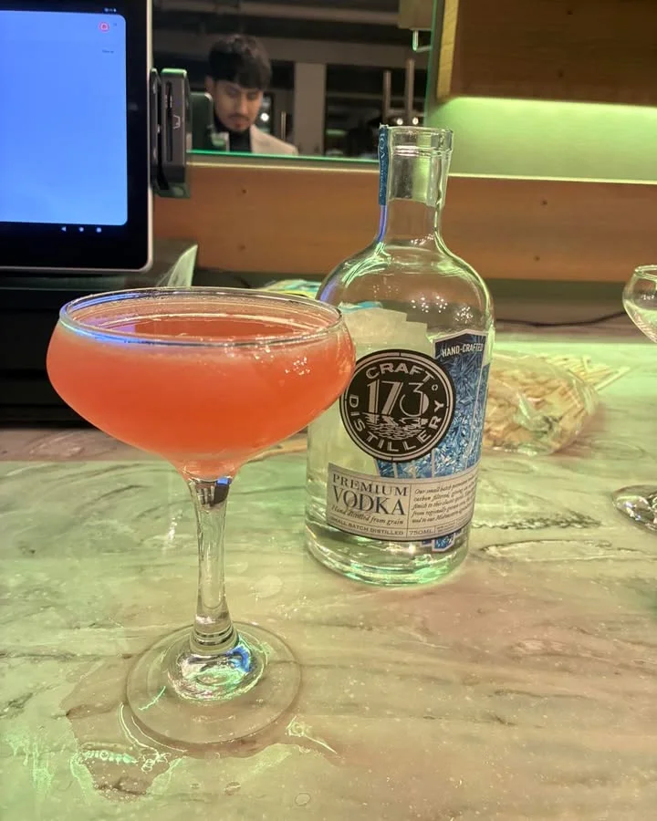 A vibrant cocktail served in a coupe glass, alongside a bottle of premium vodka on a bar table.