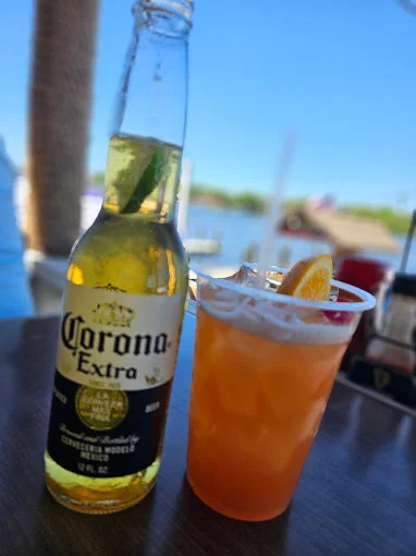 A bottle of Corona Extra next to a cocktail garnished with an orange slice and a lime wedge, with a view of the water in the background.