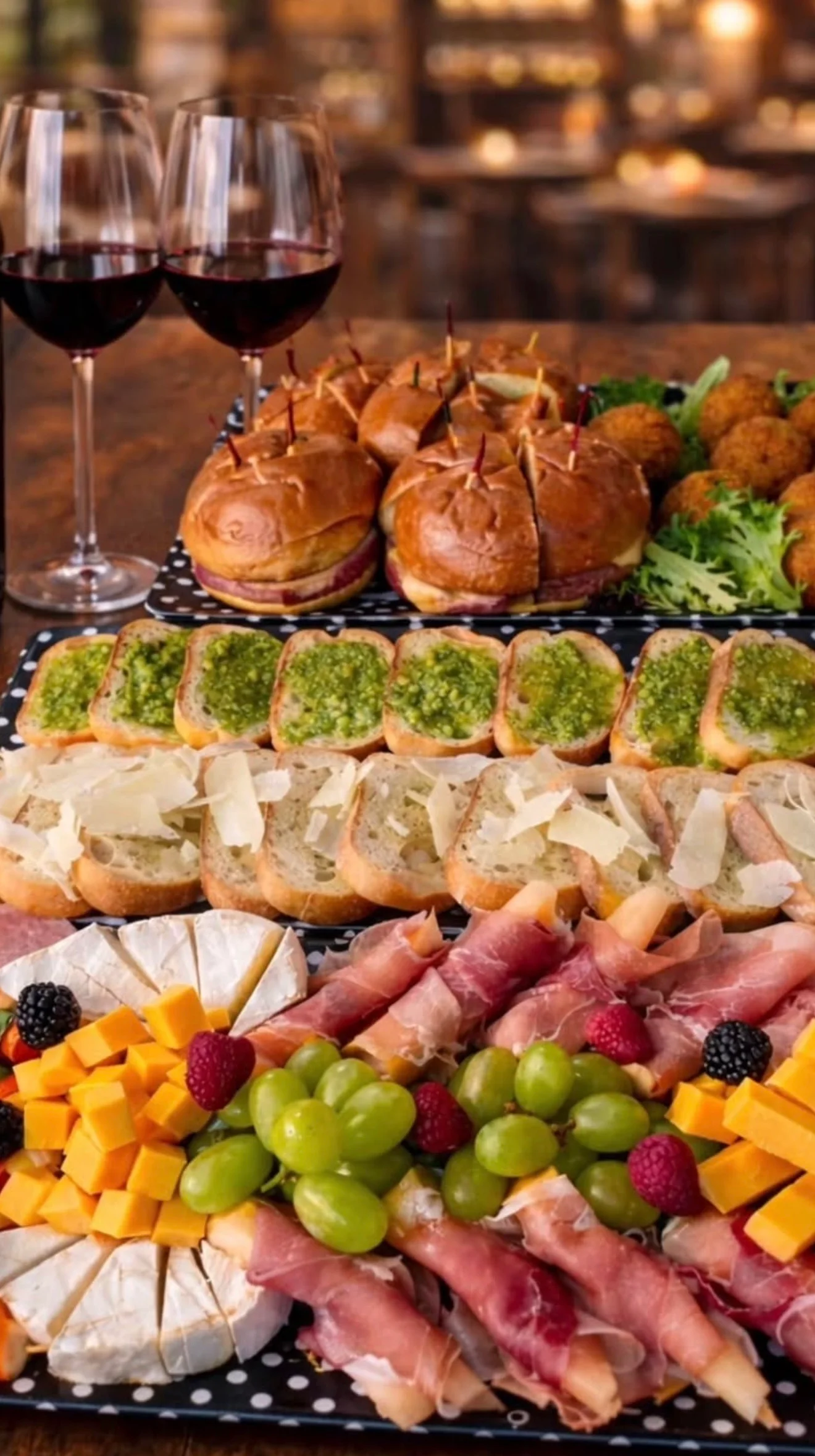 An assortment of appetizers including sliders, crostini with pesto, cheese, prosciutto, grapes, and wine glasses arranged beautifully on a wooden table.