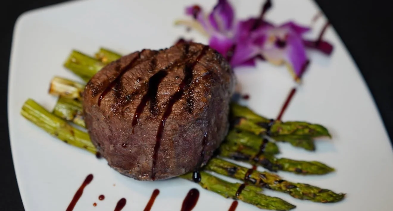 A beautifully presented filet mignon with grill marks, served on a plate with asparagus and a touch of sauce, garnished with a flower.