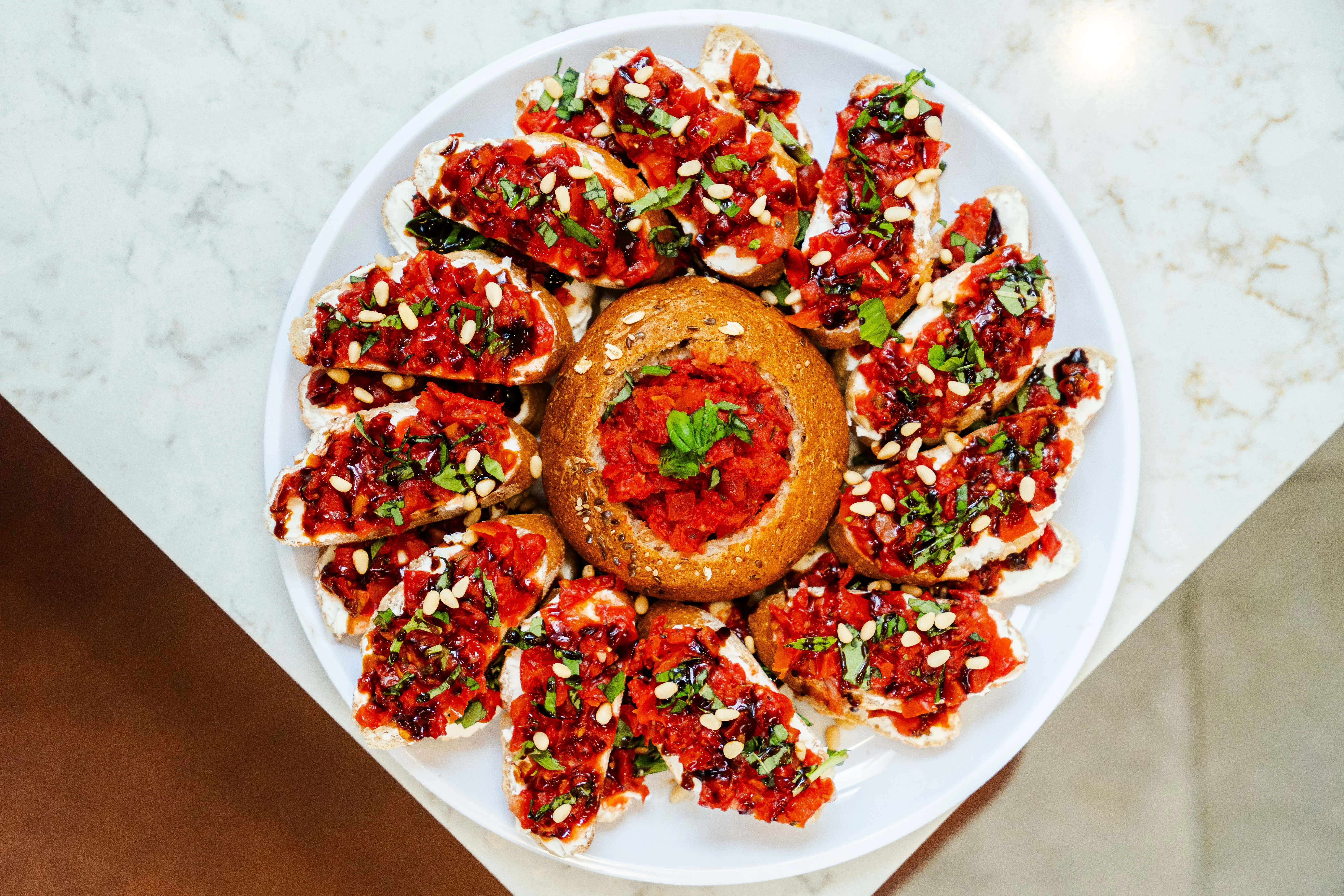 A circular platter filled with colorful toasted bread topped with roasted red peppers, herbs, and a centerpiece of bread filled with a spicy topping.
