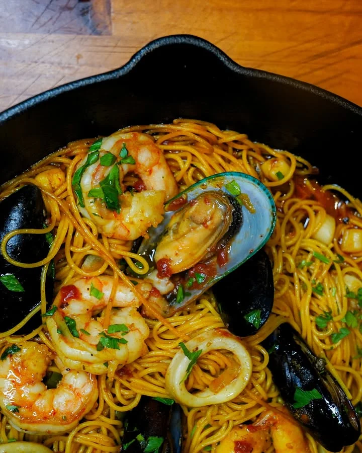 A hearty seafood pasta dish featuring shrimp, mussels, and calamari, garnished with fresh herbs on a wooden surface.