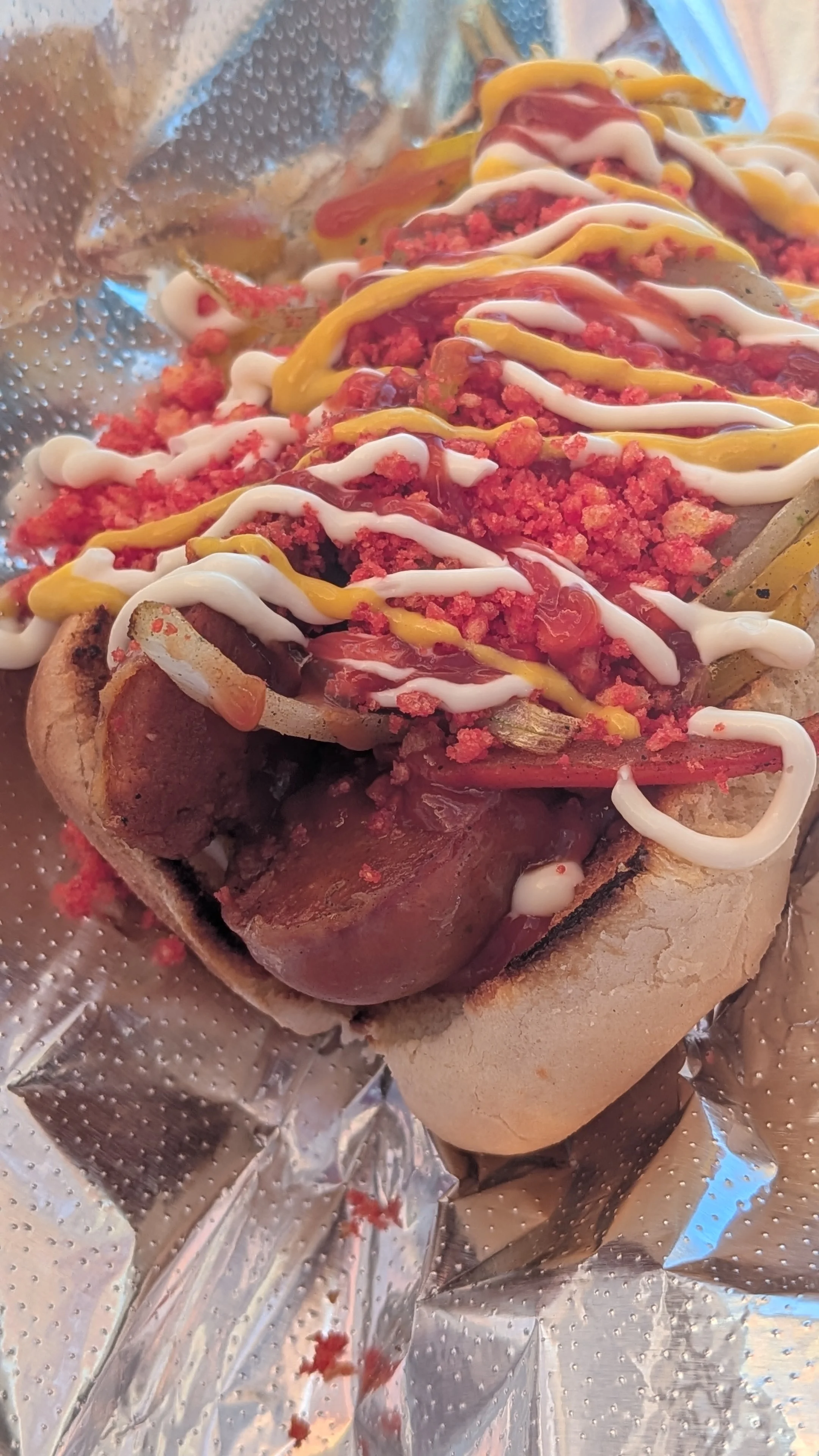 A grilled hot dog topped with a variety of condiments, including ketchup, mustard, and crushed snacks, served in a bun.