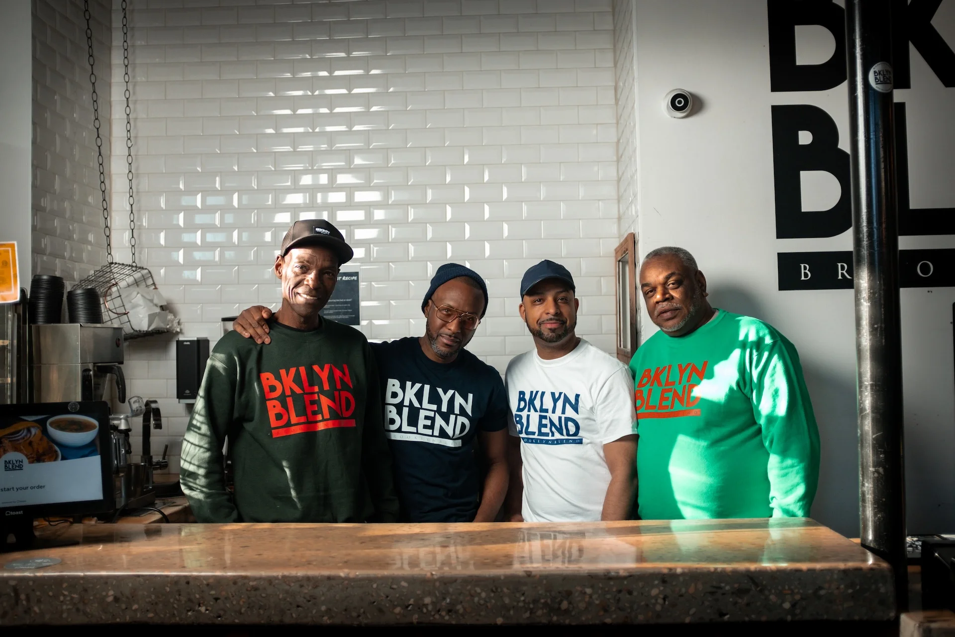 Four men wearing branded "BKLYN BLEND" shirts pose together in a cafe with a tiled wall. They are smiling, conveying a friendly and welcoming atmosphere.