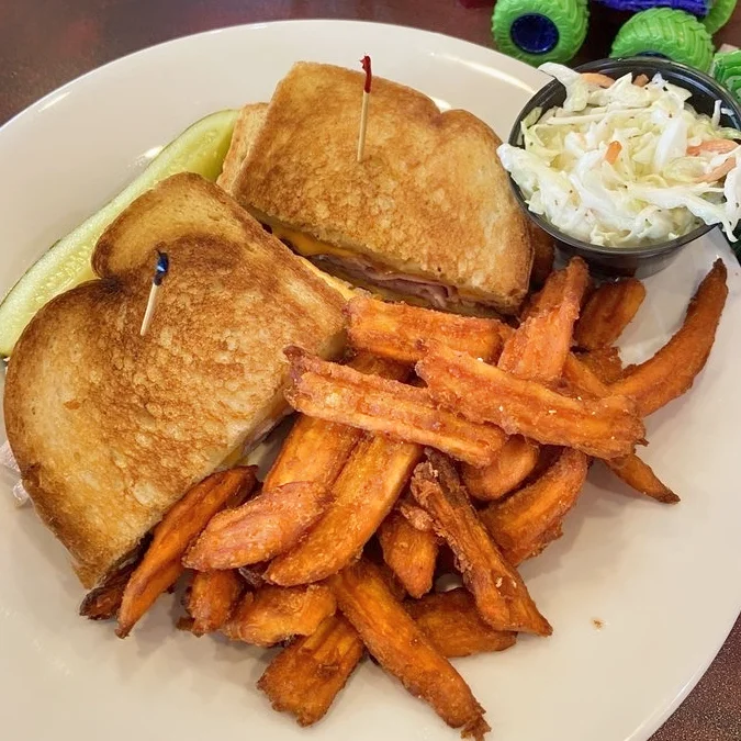 A plate featuring a grilled sandwich with melted cheese and ham, served with a side of crispy sweet potato fries, a pickle spear, and a small cup of coleslaw.