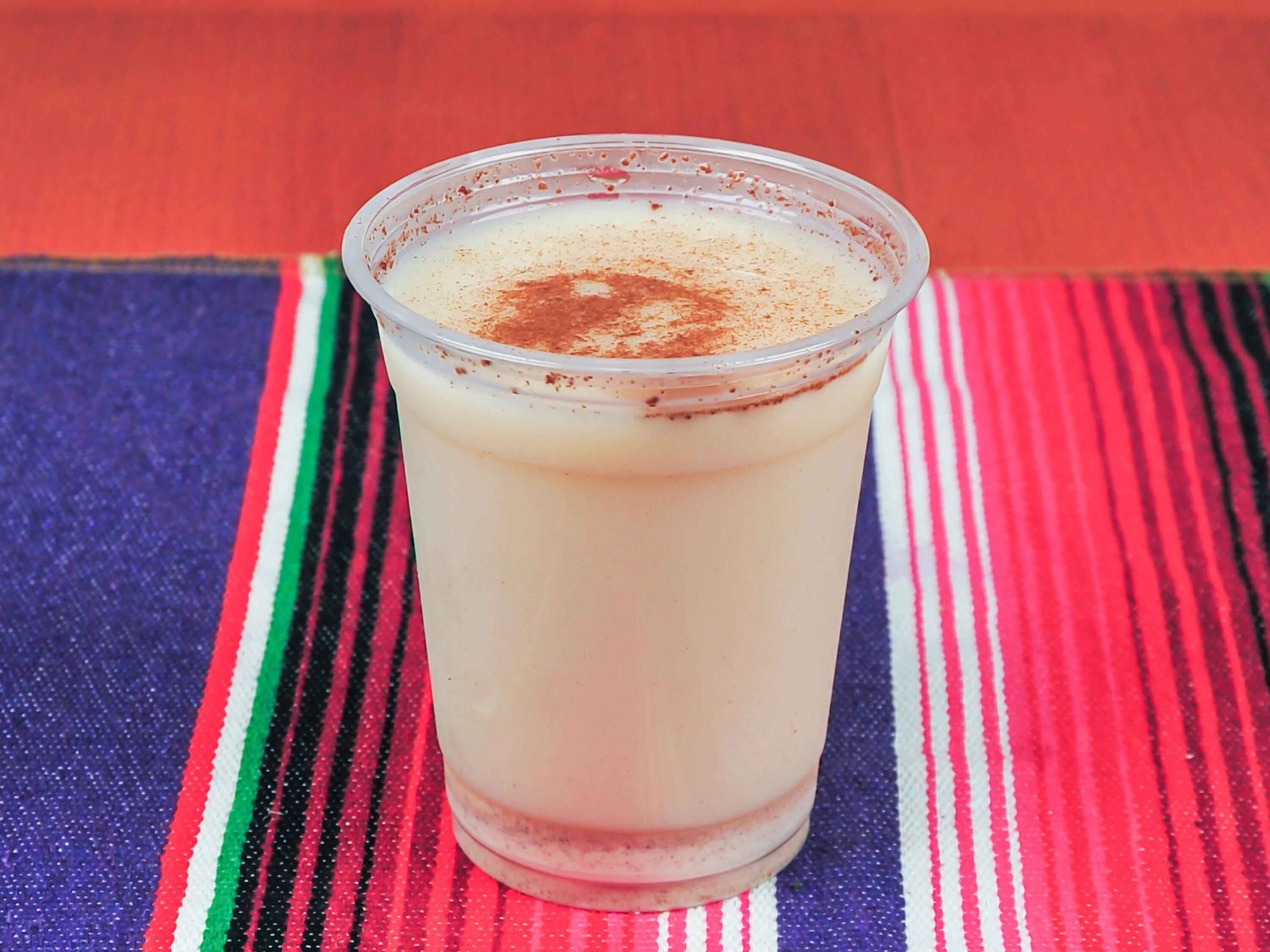 A plastic cup filled with a creamy, light-colored drink topped with a sprinkle of cinnamon. The cup is set on a vibrant striped tablecloth.
