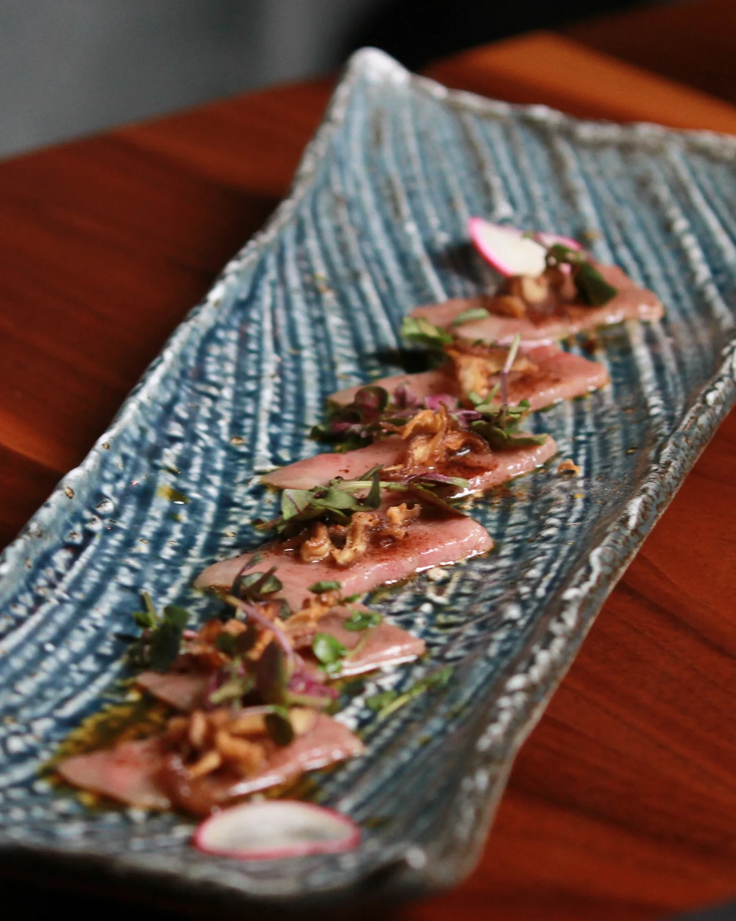 Slices of seared beef tataki or tuna carpaccio arranged in a row down a long, textured blue ceramic plate, garnished with microgreens, crispy fried shallots, and thin slices of radish on a wooden table.
