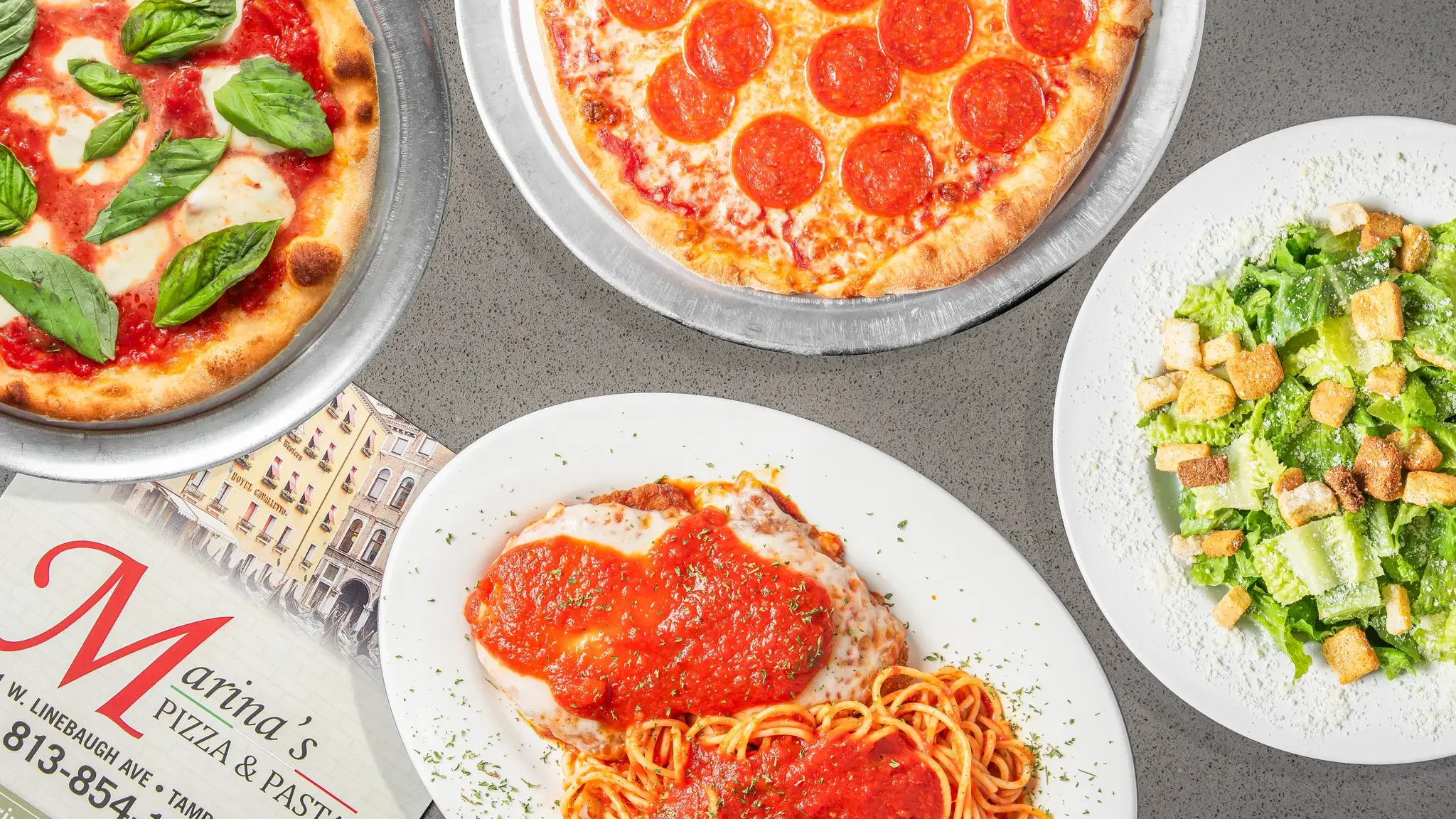 A variety of Italian dishes is laid out on a wooden table, featuring a large pizza topped with vegetables and meat, a plate of spaghetti with meat sauce and chicken parmesan, a cheesy calzone, and a cheesesteak sandwich.