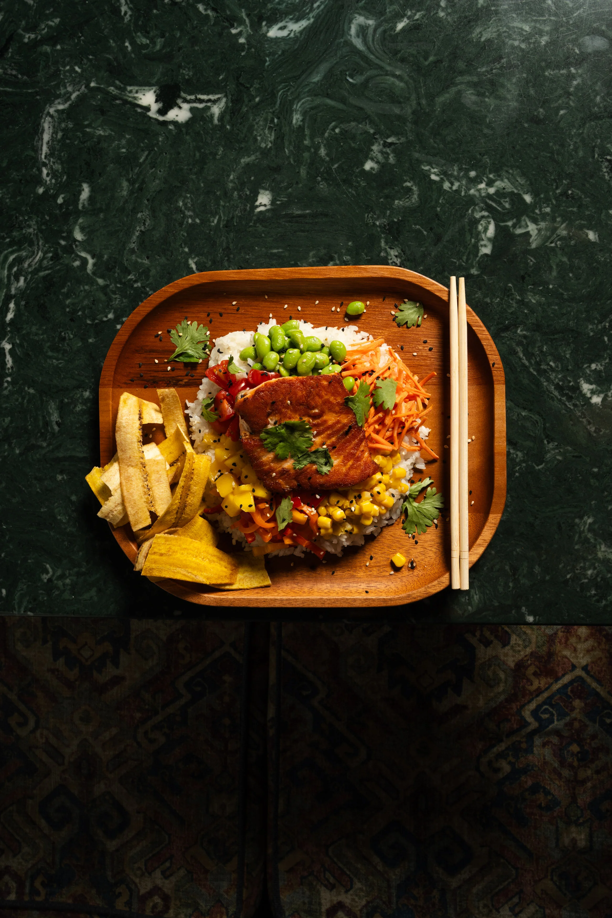 A wooden platter filled with a colorful rice bowl topped with salmon, vegetables, and garnished with cilantro, accompanied by crispy plantain chips.