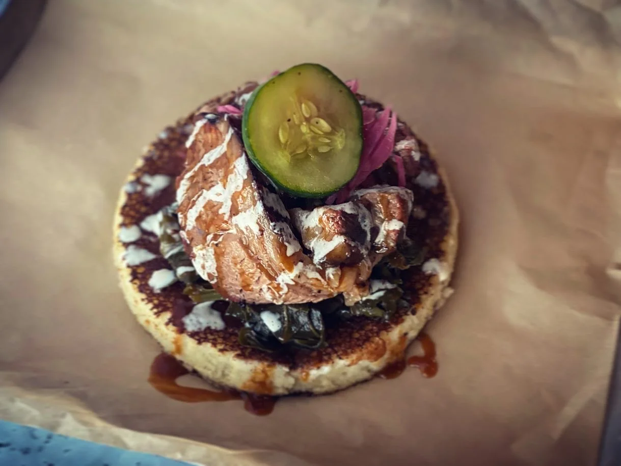 A gourmet taco topped with salmon, pickled onions, cucumbers, and a drizzle of sauce, served on a toasted corn tortilla.