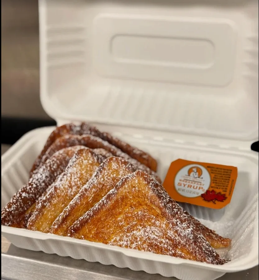 A takeout container filled with golden-brown French toast slices and a side of syrup. The slices are dusted with powdered sugar.