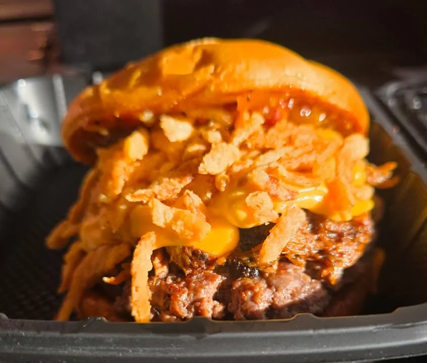 A savory hamburger topped with melted cheese and crispy onion strings, presented in a takeout container.