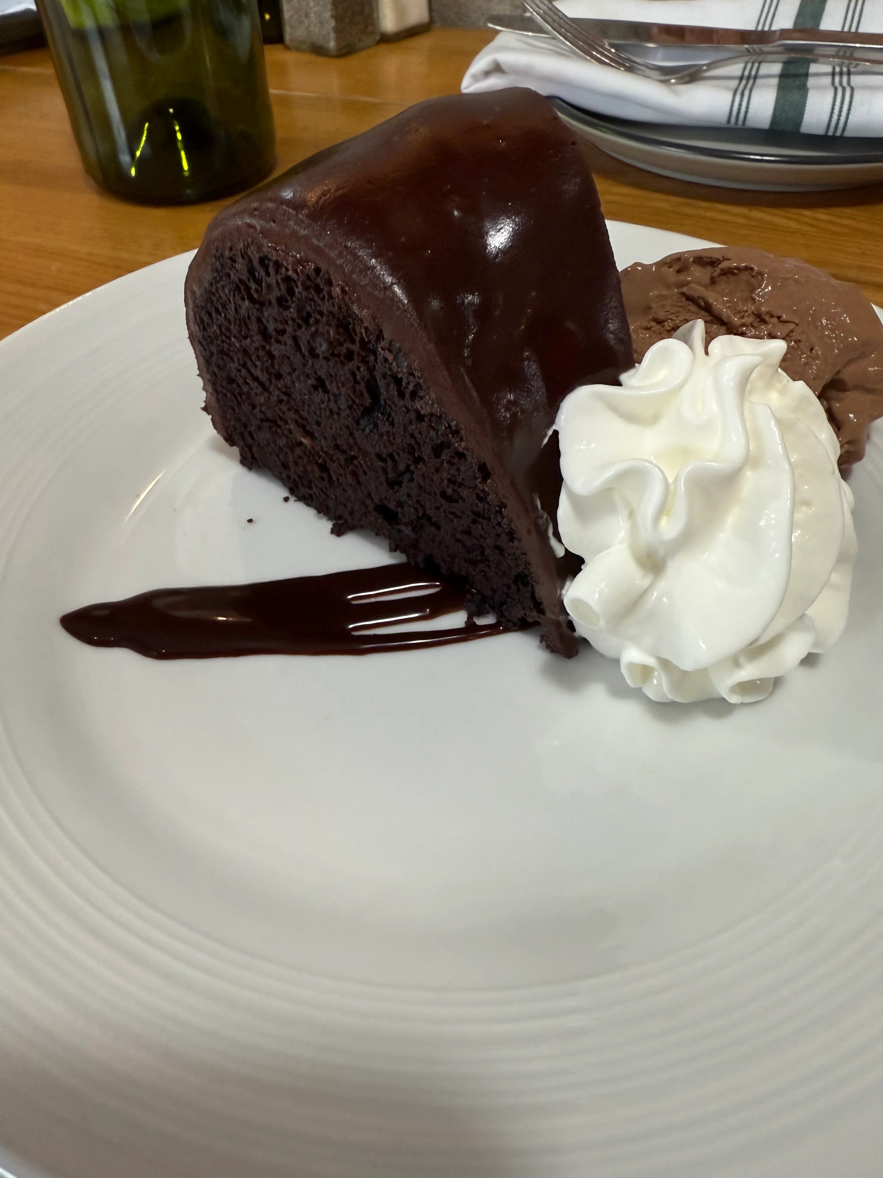A slice of chocolate cake with a glossy chocolate glaze, accompanied by whipped cream and a scoop of chocolate ice cream on a white plate.