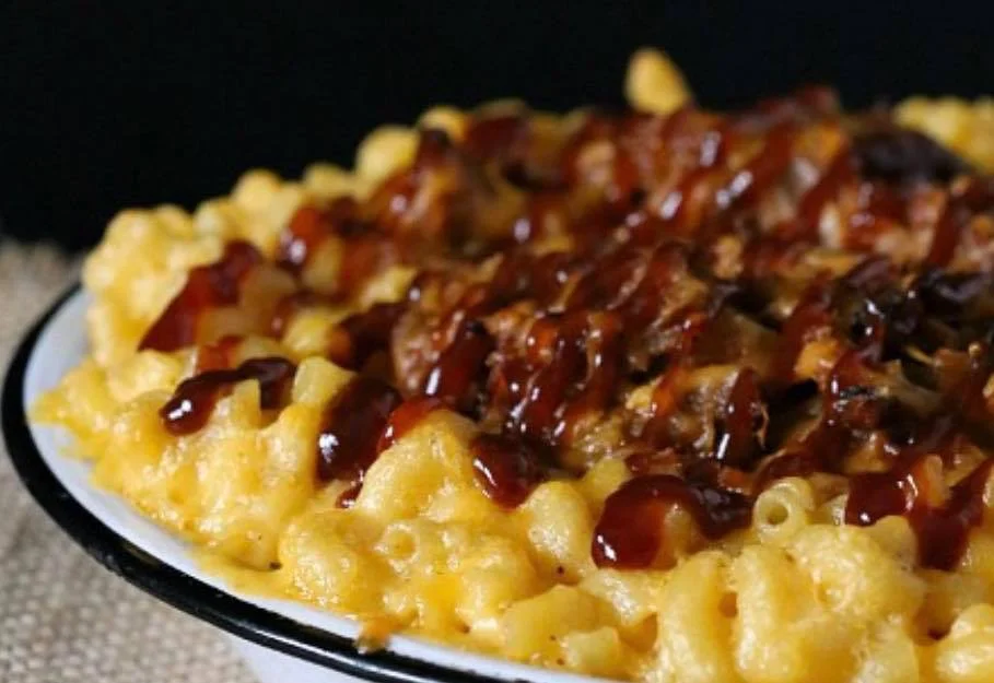 A dish of creamy macaroni and cheese topped with shredded meat and drizzled with barbecue sauce.