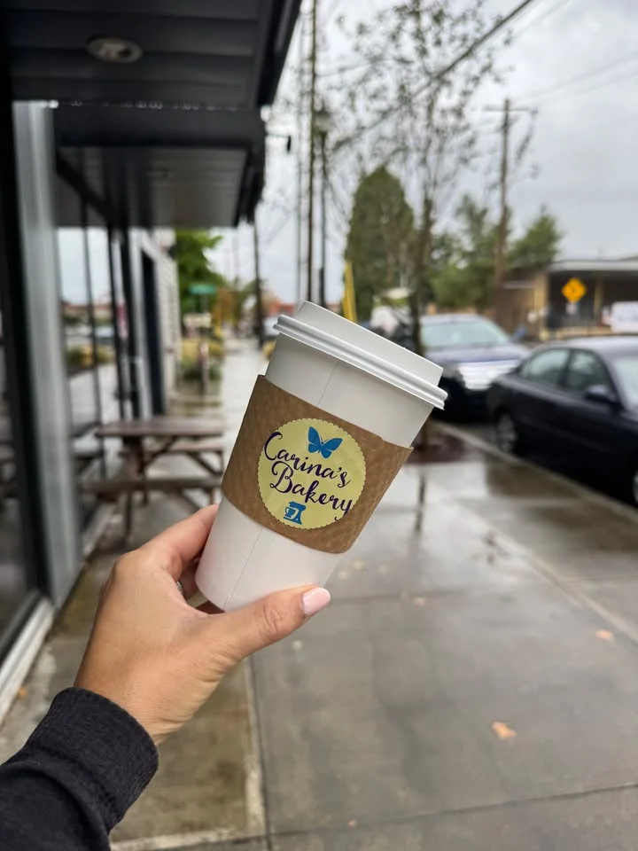 A person holding a coffee cup with a logo from a bakery. Raindrops are visible on the ground, indicating the weather is rainy.