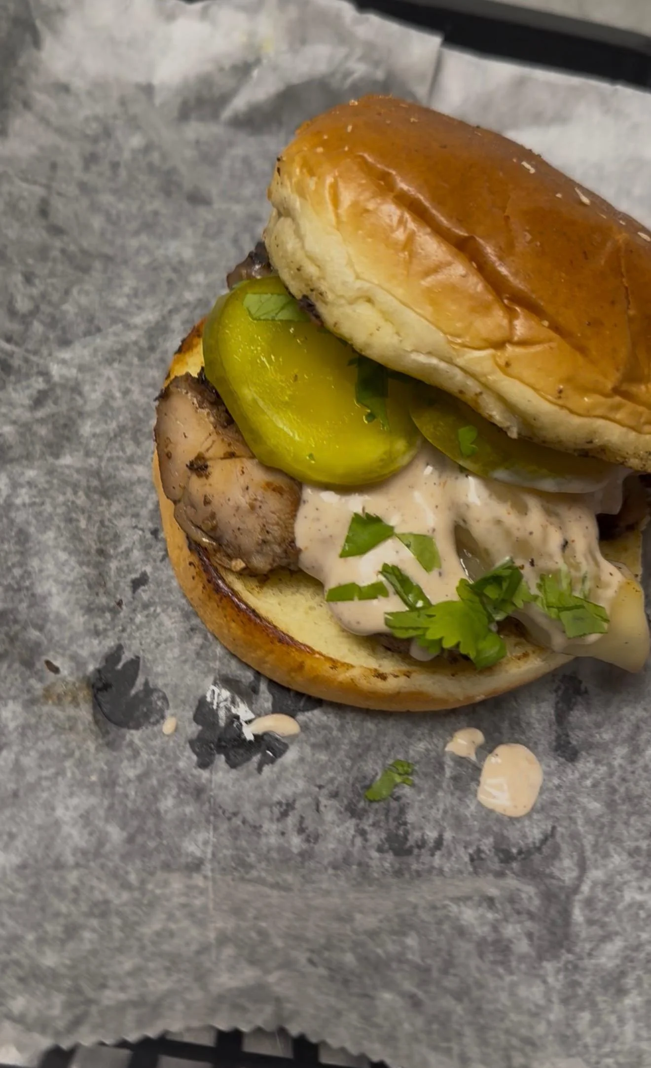 A sandwich featuring grilled chicken, pickles, and cilantro, served on a toasted bun with a creamy sauce.