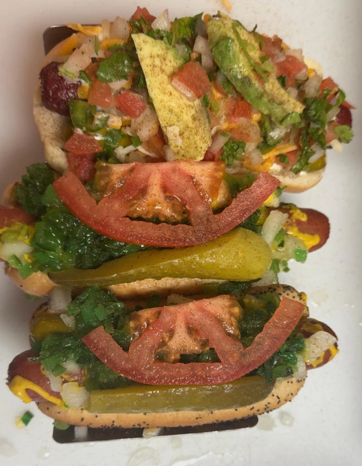 Three loaded hot dogs topped with diced vegetables, avocado, and condiments, served on a white plate.